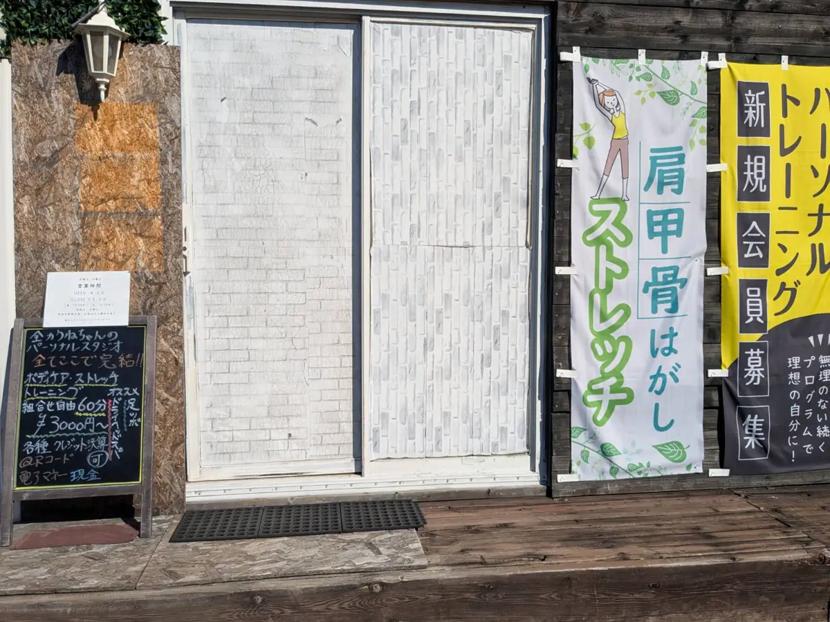 スタジオの入口。白い引き戸の横に黒板の看板と「肩甲骨はがしストレッチ」「パーソナルトレーニング 新規会員募集」ののぼり