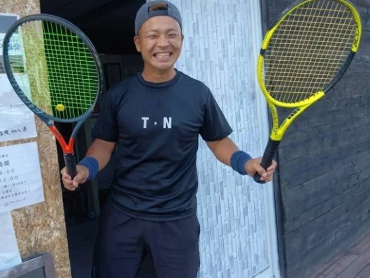 代表 采本智幸。スタジオ前でテニスラケットを両手に持って笑顔